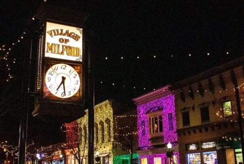 A clock tower sign reading "Village of Milford" illuminated at night, surrounded by colorful holiday lights on buildings.