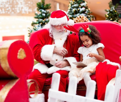 A child in reindeer antlers sits with Santa Claus, looking at a tablet, surrounded by Christmas trees.