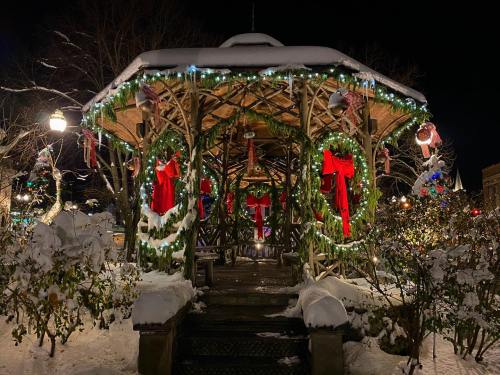 A festive gazebo adorned with wreaths and bows, surrounded by snow and holiday lights at night.