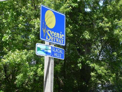 A blue sign for a scenic byway with a sun graphic, pointing to "Glacial Hills" below it, surrounded by green trees.