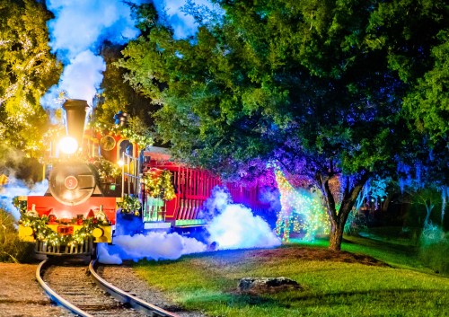 A brightly lit train decorated with festive lights, surrounded by trees and smoke, creating a magical nighttime scene.