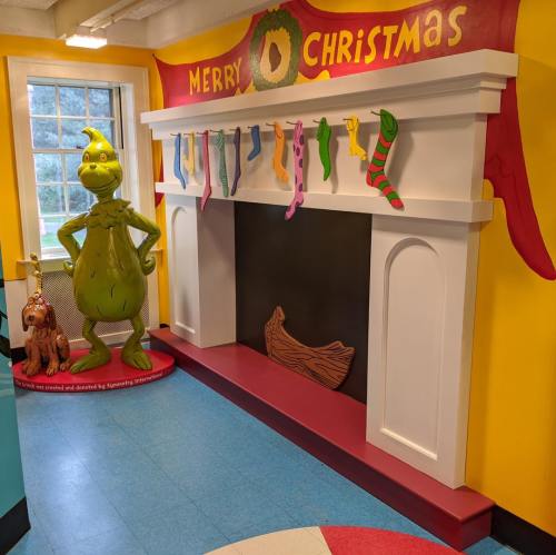 A colorful Christmas-themed room featuring a Grinch statue beside a decorated fireplace and stockings.