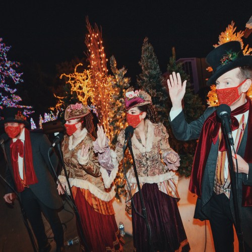 A group of performers in festive attire singing outdoors, surrounded by twinkling lights and decorated trees.