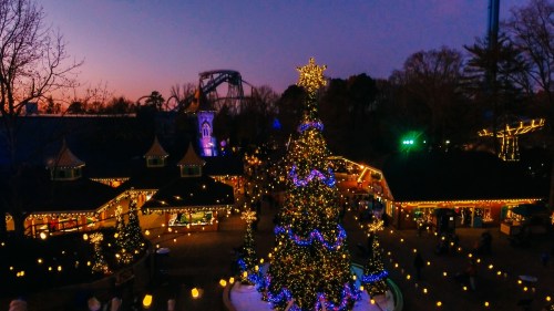 A beautifully lit Christmas tree surrounded by festive decorations and lights at dusk in a theme park.