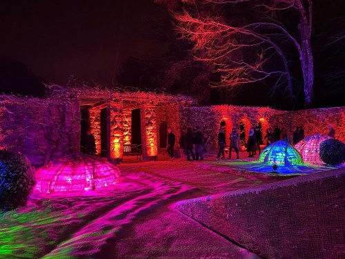 Colorful lights illuminate a stone structure and igloo-like installations in a snowy garden at night.