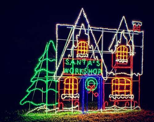 Colorful Christmas lights form a festive display of Santa's workshop with a tree beside it.