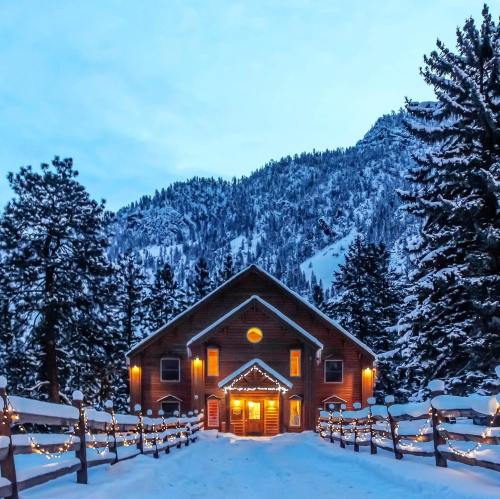 A cozy cabin adorned with lights, surrounded by snow-covered trees and mountains under a twilight sky.