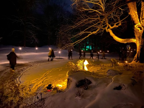 A snowy path at night, illuminated by torches, with people walking and a glowing fire in the foreground.