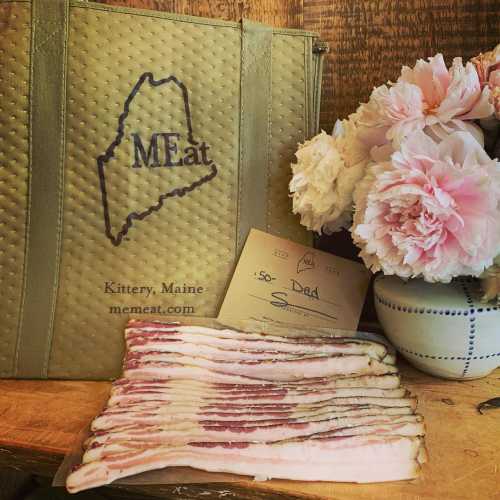 A wooden table with a Maine-themed bag, fresh flowers, and a plate of bacon strips.