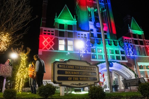 A couple stands near a festive sign in front of a brightly lit building decorated for the holidays.