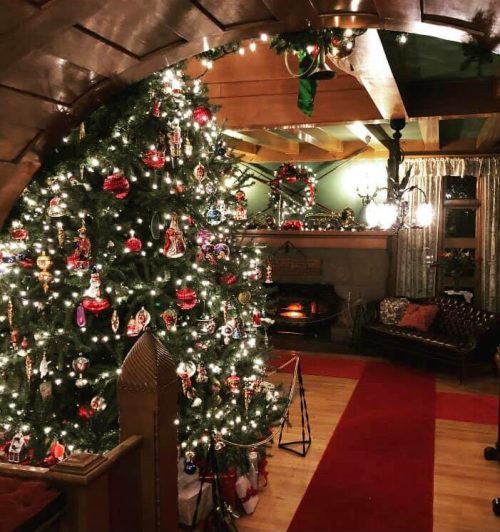 A cozy room decorated for Christmas, featuring a brightly lit tree, ornaments, and a warm fireplace.