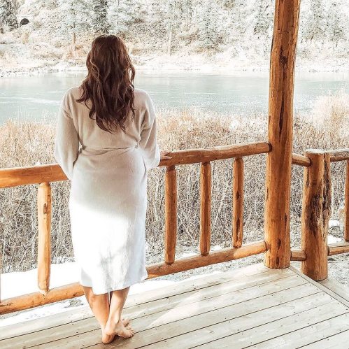 A person in a white robe stands on a wooden deck, overlooking a serene river and snowy landscape.