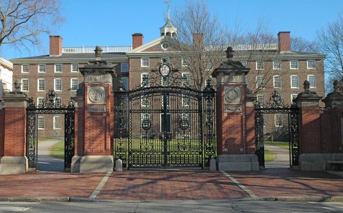 Iron gates with brick pillars lead to a historic building on a sunny day, surrounded by trees and a clear blue sky.