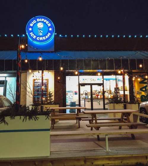 Exterior of Big Dipper Ice Cream shop at night, featuring bright signage and outdoor seating with festive lights.
