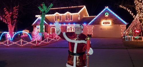 A man dressed as Santa waves in front of a brightly lit house decorated for Christmas.