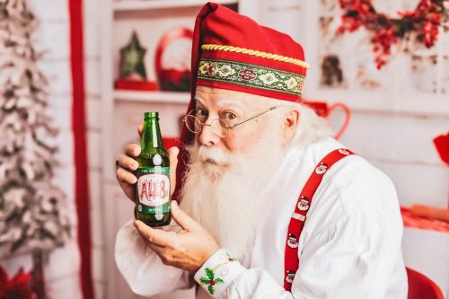 Santa Claus in festive attire holds a green bottle labeled "Ale 8" with a cheerful expression in a holiday setting.