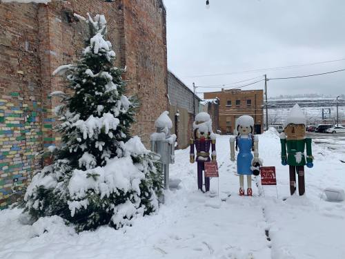 Snow-covered scene featuring a decorated tree and wooden figures in winter attire, set against a brick wall.