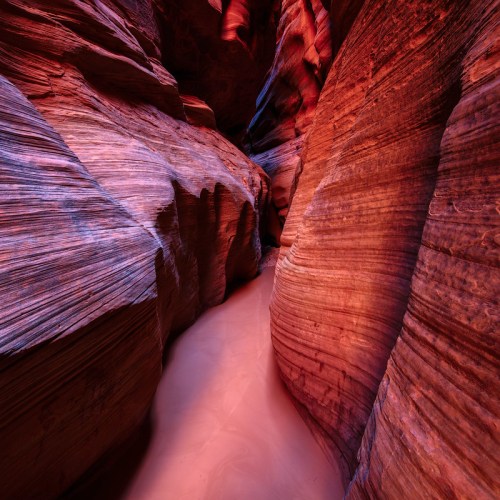 A narrow canyon with vibrant red and orange rock formations and soft sand at the bottom, illuminated by natural light.