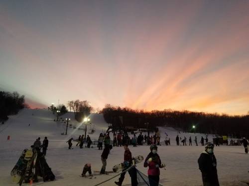Skiers and snowboarders on a snowy slope at sunset, with vibrant colors in the sky and trees in the background.