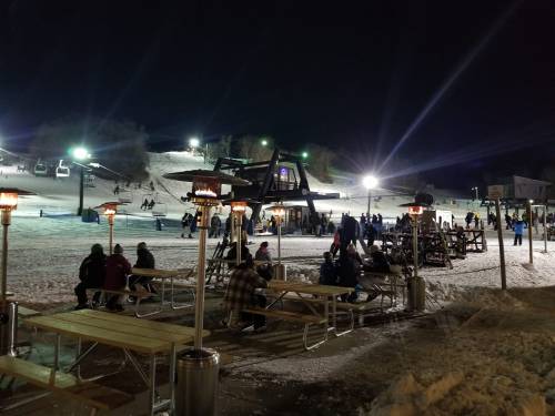 A snowy ski resort at night, with people gathered around tables and ski lifts illuminated in the background.