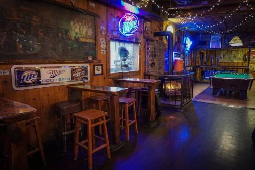 Cozy bar interior with wooden tables, stools, neon signs, and a pool table, illuminated by warm lights.
