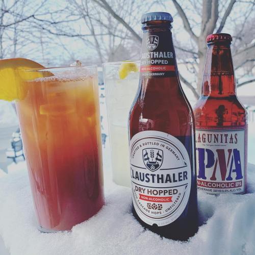 Three beverages on snow: a cocktail with an orange slice, and two non-alcoholic beer bottles.