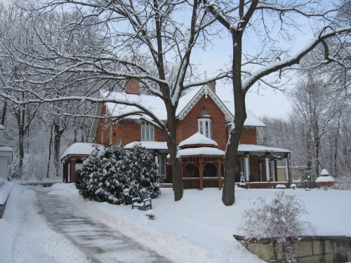 A snow-covered house surrounded by trees, with a path leading up to it and a winter landscape in the background.