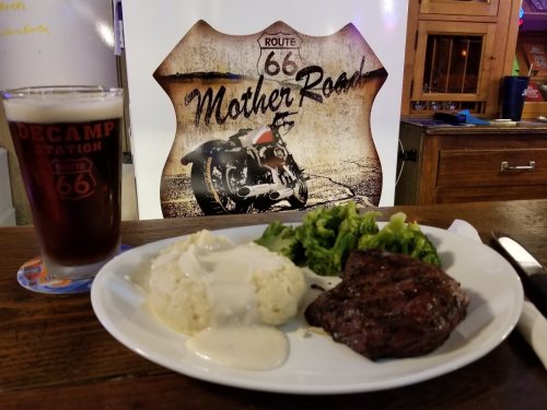 A plate with steak, mashed potatoes with gravy, and broccoli, alongside a pint of beer, with a Route 66 sign in the background.