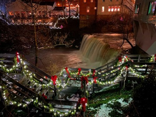 A festive scene featuring a waterfall surrounded by holiday lights and decorations, with a cozy town in the background.