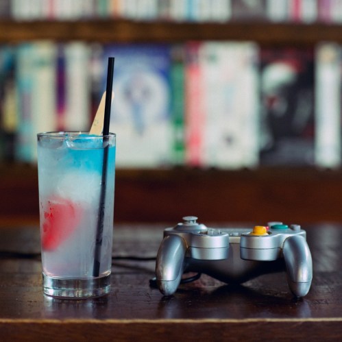A colorful cocktail with a straw next to a video game controller on a wooden table, with books in the background.