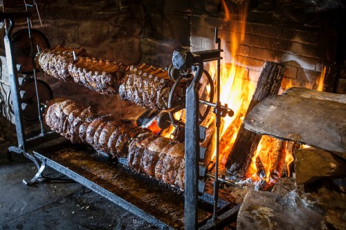 A rustic fireplace with a spit roasting several cuts of meat over an open flame, creating a warm, inviting atmosphere.