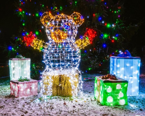 A lit teddy bear made of lights stands among colorful gift boxes in a snowy outdoor setting.