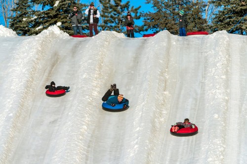A snowy hill with people tubing down, while others watch from the top, surrounded by trees and a clear blue sky.