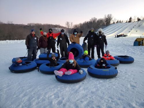 A group of people in winter attire poses on snow tubes, with a snowy hill in the background.