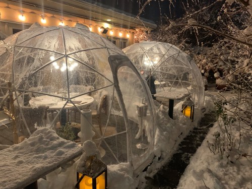 Two transparent igloos in a snowy outdoor setting, illuminated by warm lights, with tables inside.