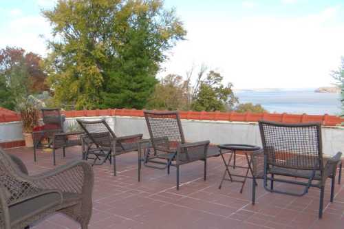 A scenic outdoor patio with several chairs and a small table, overlooking a body of water and trees in the background.