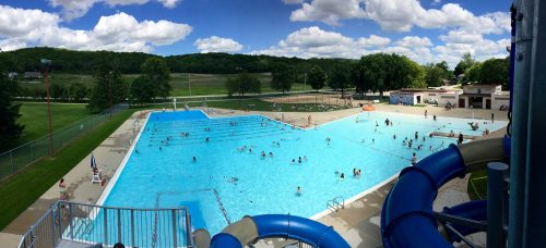 A large outdoor swimming pool filled with people, surrounded by green trees and a blue sky with fluffy clouds.