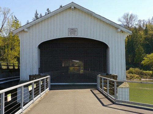 A white covered bridge with a wooden structure, leading to a pathway over a river, surrounded by trees.