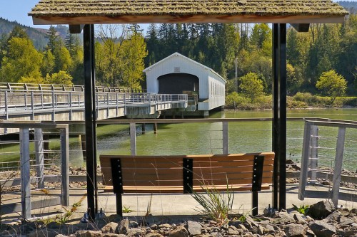 A wooden bench under a shelter overlooks a calm river and a white covered bridge in a scenic, green landscape.