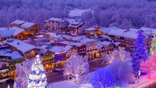 A snowy village scene at dusk, with colorful lights on trees and buildings, creating a festive winter atmosphere.