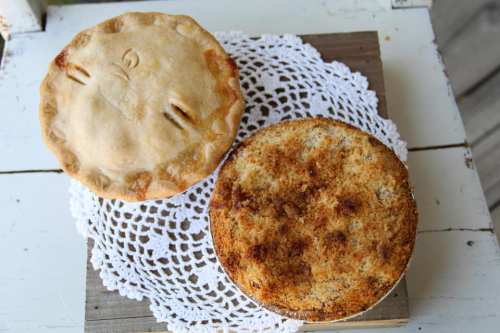 Two baked pies on a lace doily: one is a golden-brown pie with a decorative crust, the other is a crisp, crumb-topped pie.