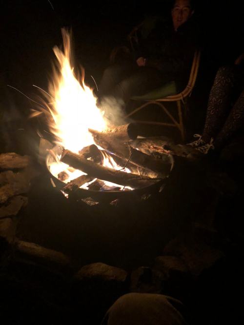 A campfire burns brightly in a stone fire pit, with people sitting nearby in the dark.