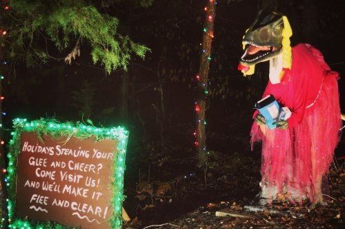 A whimsical creature in a red outfit stands by a sign with festive lights, inviting visitors to enjoy the holidays.