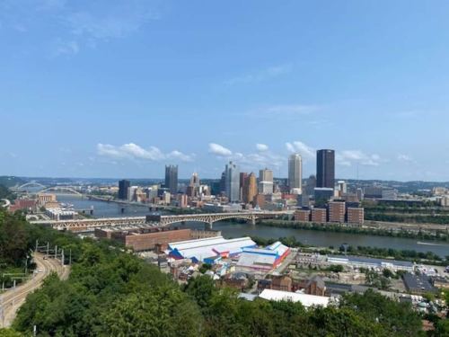 A panoramic view of Pittsburgh's skyline, featuring tall buildings and a river, under a clear blue sky.
