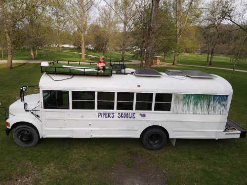 A converted school bus named "Piper's Skoolie" parked in a grassy area, with solar panels on the roof and a person sitting on top.