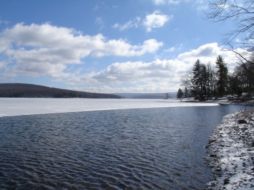 A serene winter landscape featuring a partially frozen lake, blue sky, and scattered clouds, bordered by trees.