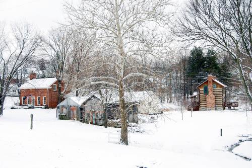 A snowy landscape featuring a brick house, wooden shed, and log cabin surrounded by bare trees.