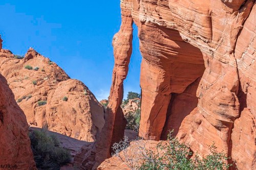 Red rock formations with a narrow arch, surrounded by desert vegetation and a clear blue sky.