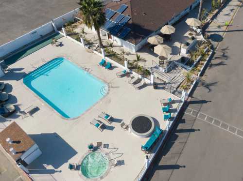 Aerial view of a pool area with lounge chairs, umbrellas, and a hot tub surrounded by palm trees and a building.