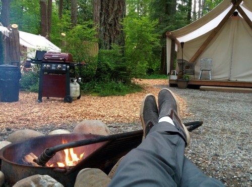 A person relaxes by a campfire, with a tent and grill visible in a forested campsite setting.